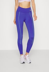 Leggings viola con strisce laterali verdi, vita alta e tessuto elasticizzato. Indossati con scarpe da ginnastica chiare. Texture liscia.