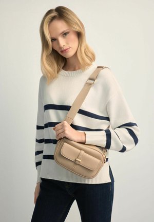 Blondynka w białym swetrze w granatowe paski i ciemnych dżinsach, trzyma beżową torebkę na ramię z szerokim paskiem przewieszonym przez klatkę piersiową.