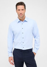 Lichtblauw lange mouwen overhemd met een button-down kraag, voorzien van zwarte knopen en een zachte textuur. Draagt bij donkere marineblauwe broek.