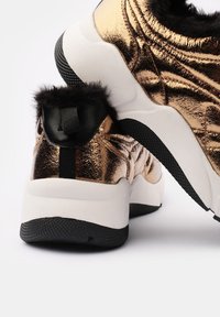 Sneaker metallici oro e neri con una superficie testurizzata, dettagli in pelliccia sintetica sul tallone e una prominente intersuola bianca.