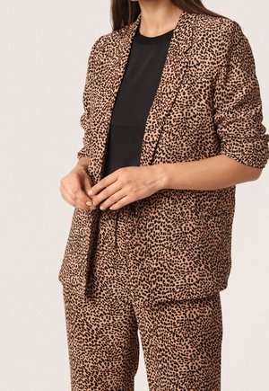 Kobieta ubrana w brązowo-czarną marynarkę w panterkę oraz pasujące spodnie, z czarną koszulą, dłonie poprawiające przód marynarki.