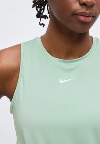 Γυναίκα που φοράει αμάνικη αθλητική μπλούζα ανοιχτού πράσινου χρώματος με λευκό λογότυπο της Nike στο κέντρο κάτω από το λαιμό, το οποίο κόβεται κάτω από το στόμα.