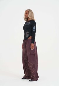 Kobieta z blond włosami ubrana w czarny, długi rękaw bodysuit z graficznymi nadrukami oraz luźne, bordowe spodnie cargo z wzorami, stojąca w profilu.
