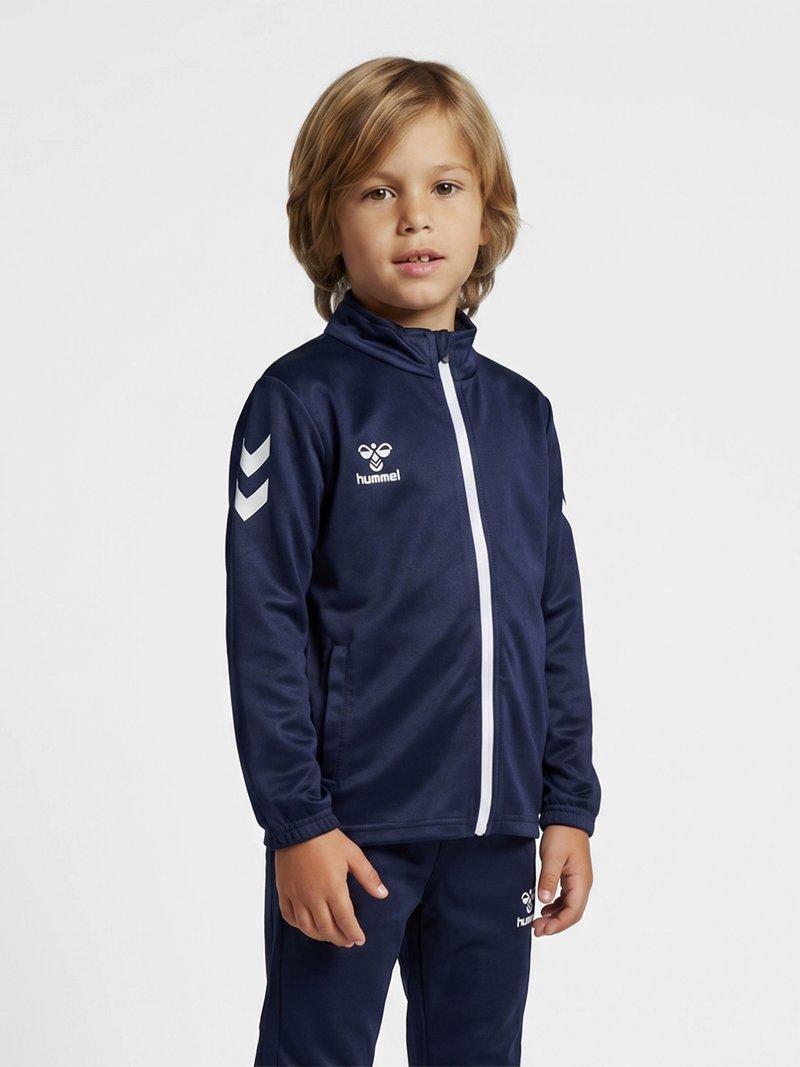 Enfant debout portant un survêtement de sport bleu marine avec une fermeture éclair blanche et des logos sur la poitrine et les manches sur fond gris.