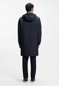 Hombre de pie de espaldas, vestido con un abrigo largo con capucha azul marino, pantalones negros y zapatillas negras con suelas blancas sobre un fondo blanco.