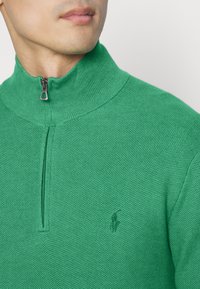 Grön halvmanschettpullover med strukturerad stickad tyg, hög krage och broderad logotypdetalj p å bröstet. Dragkedja i silverfärgad metall.