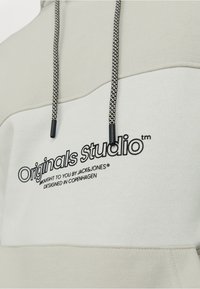 Sudadera beige clara hecha de una mezcla de algodón, con un diseño estructurado, cordones negros trenzados y el logo "Originals Studio" en el pecho.