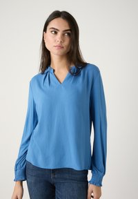 Helleblaue Bluse mit langen Ärmeln und V-Ausschnitt, weichem Stoff und gerafften Manschetten. Das Material ist glatt und leicht, ideal zum Layering.