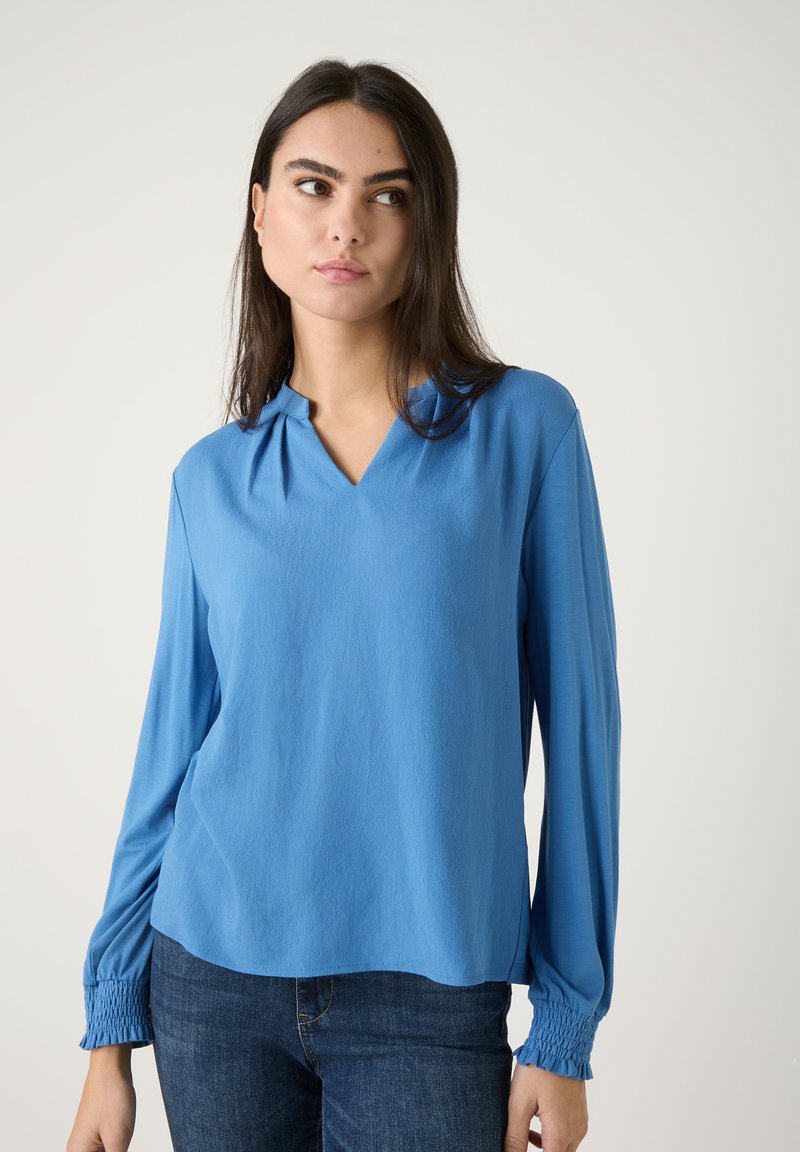 Helleblaue Bluse mit langen Ärmeln und V-Ausschnitt, weichem Stoff und gerafften Manschetten. Das Material ist glatt und leicht, ideal zum Layering.