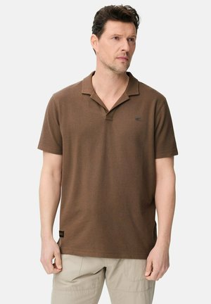 Mężczyzna ubrany w brązową koszulkę polo z krótkim rękawem i beżowe spodnie, stojący z rozluźnionymi ramionami i patrzący w bok na jednolitym tle.