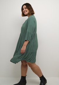 Vestido verde a la altura de la rodilla con mangas de tres cuartos, que presenta rayas verticales y una falda en capas. Se lleva con botas negras hasta el tobillo.