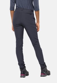 Pantaloni da esterno grigi scuri con vestibilità slim, dotati di una tasca posteriore con zip e una cintura elastica, abbinati a scarponi da trekking neri.