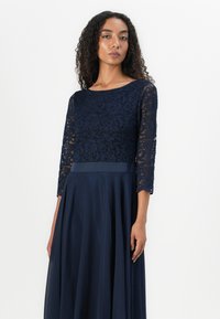 Robe de soirée bleu marine avec un corsage en dentelle à motifs floraux, des manches trois-quarts et une jupe fluide. Tissu lisse avec une ceinture en satin.