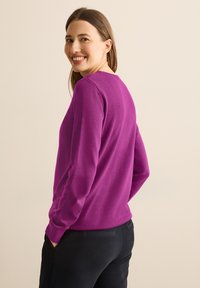 Pull à manches longues violet avec un col rond, fabriqué en matériau doux, associé à un pantalon noir texturé. Design simple sans motifs visibles.
