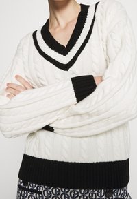 Bílý svetr s kabelovým vzorem, černý proužkovaný výstřih do V a manžety. Má žebrovaný lem a rukávy, které zdůrazňují strukturu a kontrast.