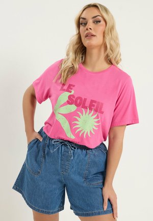 Blonde Frau trägt ein pinkfarbenes T-Shirt mit dem Schriftzug "LE SOLEIL" und Grafik, kombiniert mit lockeren Jeansshorts, posiert mit der Hand in der Tasche.