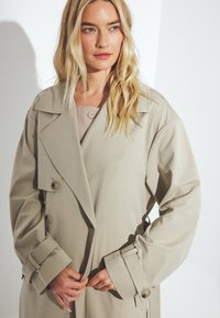 Bežs trenčkotis ar dubultu pogu dizainu, lielu apkakli un pogu detaļām. Audums izskatās gluds ar nedaudz strukturētu piegriezumu.