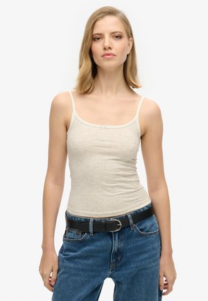 Joven mujer con una camiseta de tirantes beige y vaqueros azules con cinturón negro, de pie frente a un fondo blanco liso.