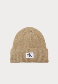 Gorro de punto beige con un dobladillo, que presenta una etiqueta rectangular blanca con el logotipo "cK" en negro. Textura suave y patrón de canalé en todo el gorro.