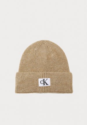 Berretto lavorato a maglia beige con risvolto, caratterizzato da un'etichetta rettangolare bianca con il logo "cK" nero. Texture morbida e motivo a coste in tutto il berretto.