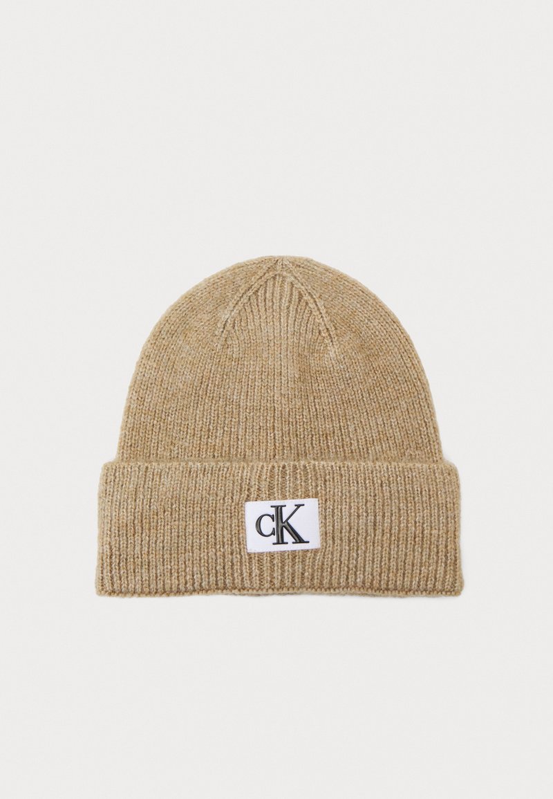 Gorro de punto beige con un dobladillo, que presenta una etiqueta rectangular blanca con el logotipo "cK" en negro. Textura suave y patrón de canalé en todo el gorro.