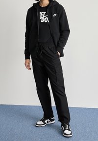 Black zip-up hoodie with kangaroo pockets, black t-shirt featuring "JUST DO IT" in white, black cargo pants, and black/white sneakers.