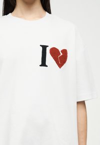 Λευκό T-shirt με μαύρο γράμμα «I» και κεντημένο υφασμάτινο κόκκινο σπασμένη καρδιά σύμβολο στην αριστερή πλευρά του στήθους.