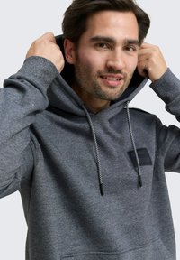 Grijze hoodie met een koord. Heeft een gestructureerde stof, een patch op de borst en zwarte accenten op de koorden en zakken.