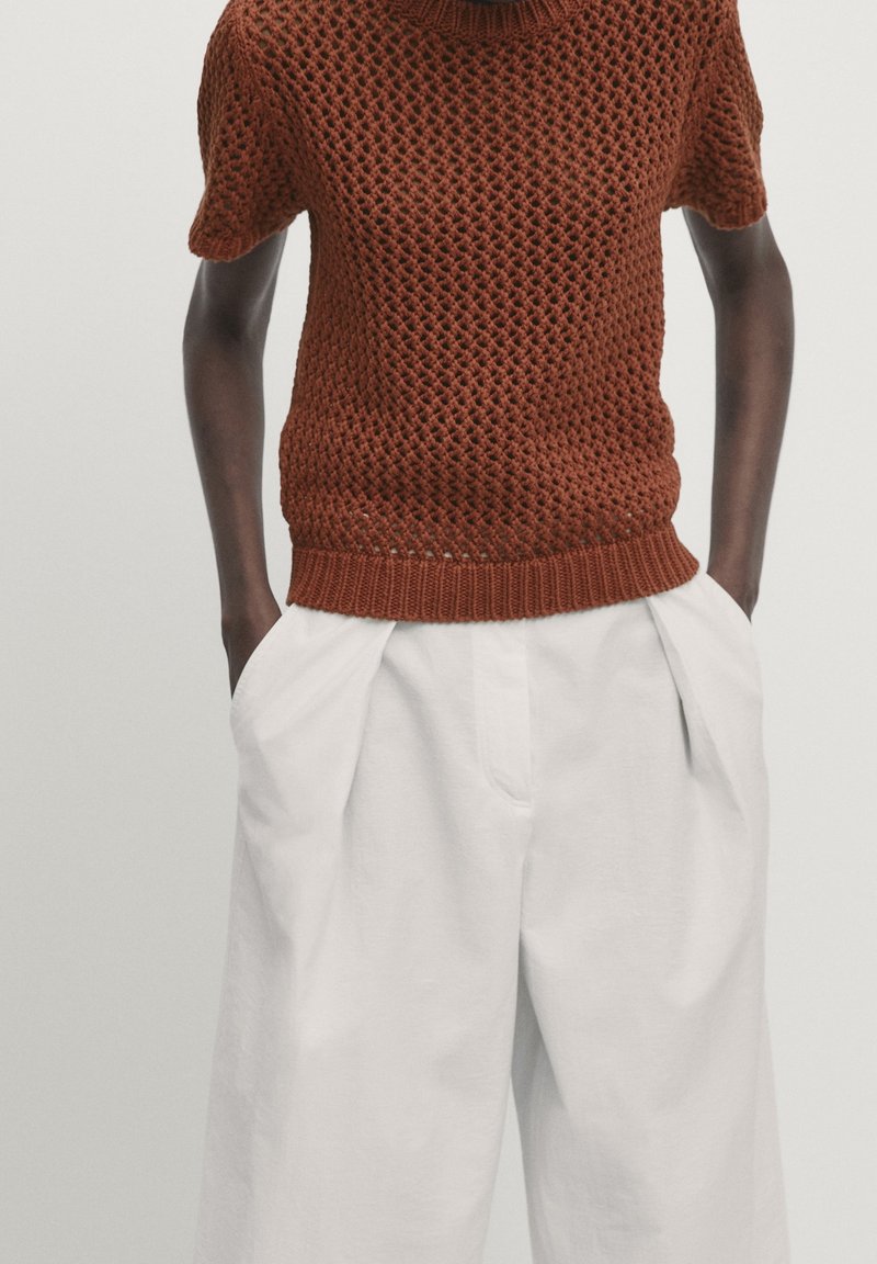 Maglione corto a maniche corte di colore marrone con un motivo testurizzato, abbinato a pantaloni bianchi a pieghe larghi.