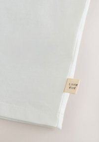 Valge puuvillane T-särk pehme tekstuuriga, mille ääres on väike beež silt "Little Bird", lihtne kujundus ja puhtad jooned.