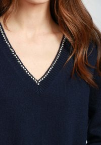 Donna con lunghi capelli castani che indossa un maglione blu navy a scollo a V con decorazioni in strass lungo il collo.