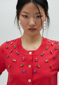 Cardigan rouge en tricot avec un col rond, des fermetures à boutons dorés et des bijoux décoratifs multicolores en perles le long des épaules.