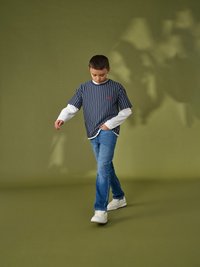 Camicia a righe blu navy e bianca a maniche corte indossata sopra una maglietta bianca a maniche lunghe, abbinata a jeans blu chiaro sbiaditi e sneakers bianche.