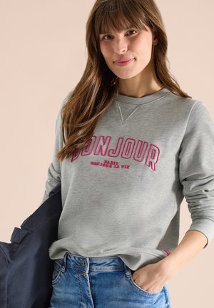 Jeune femme aux longs cheveux bruns portant un sweat-shirt gris avec le texte « BONJOUR PARIS BONJOUR LA VIE », un jean bleu et tenant un sac bleu marine.