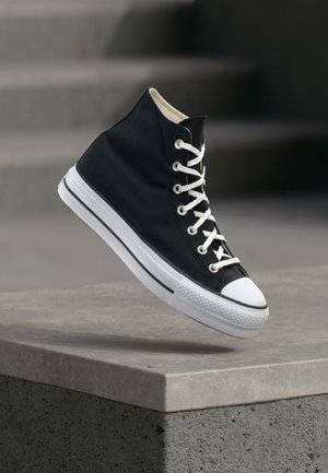Zwarte hoge sneaker met witte veters en rubberen zool, balancerend op de rand van een grijs betonnen platform.