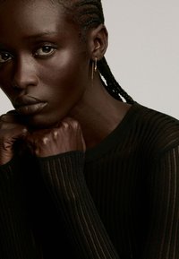Top de manga larga negro con textura acanalada. Un delicado pendiente dorado y un peinado trenzado acentúan una expresión concentrada.