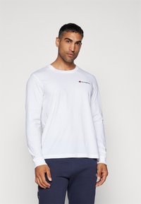 Langermet hvit bomull t-skjorte med rund hals og en liten rød og svart logo på brystet. Minimalistisk design, avslappet passform.