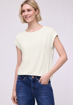 Uśmiechnięta blondynka, ubrana w kremowy top z krótkim rękawem i niebieskie dżinsy, z prawą ręką w kieszeni, na jednolitym tle.