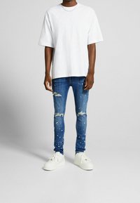 Vit bomullsoverdel i oversized modell, slitna blå jeans med färgfläckar och vita sneakers med texturerad yta och logodetalj.