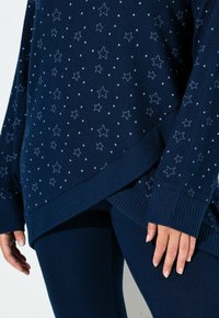 Dunkelblauer Pullover und Leggings mit weißen Sternmustern, gerippte Bündchen und Saum, getragen von einer Person mit sichtbaren Händen an den Seiten.