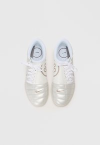 Chuteiras de futebol prateadas com um cabedal sintético brilhante, colarinho acolchoado branco, atacadores brancos e uma discreta marcação da Nike. Apresenta uma textura acolchoada.