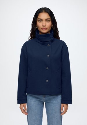 Femme aux cheveux ondulés foncés portant une veste bleu marine boutonnée avec un col montant et un jean bleu clair, debout devant un fond uni.