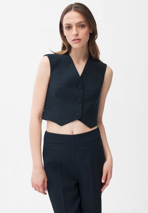 Młoda kobieta ubrana w bezrękawnik w kolorze czarnym, krótki, z trzema guzikami, oraz dopasowane, czarne spodnie z wysokim stanem, na jednolitym białym tle.