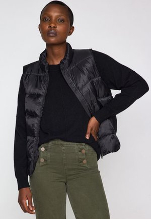 Gilet imbottito nero con colletto alto, chiusura con zip e design trapuntato, abbinato a un maglione nero e pantaloni verde oliva con dettagli a bottoni.