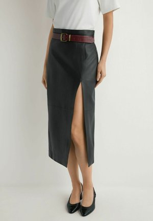 Kobieta w czarnej skórzanej midi spódnicy z przednim rozcięciem, białej bluzce z krótkim rękawem, bordowym pasku i czarnych szpilkach z ostrym noskiem.