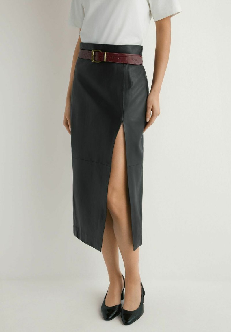 Kobieta w czarnej skórzanej midi spódnicy z przednim rozcięciem, białej bluzce z krótkim rękawem, bordowym pasku i czarnych szpilkach z ostrym noskiem.