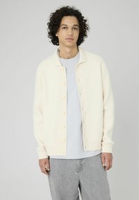 Cardigan beige clair avec une fermeture à boutons, des poignets côtelés et un tissu doux et texturé, associé à un t-shirt gris et un pantalon gris clair.