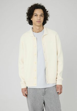 Cardigan beige clair avec une fermeture à boutons, des poignets côtelés et un tissu doux et texturé, associé à un t-shirt gris et un pantalon gris clair.