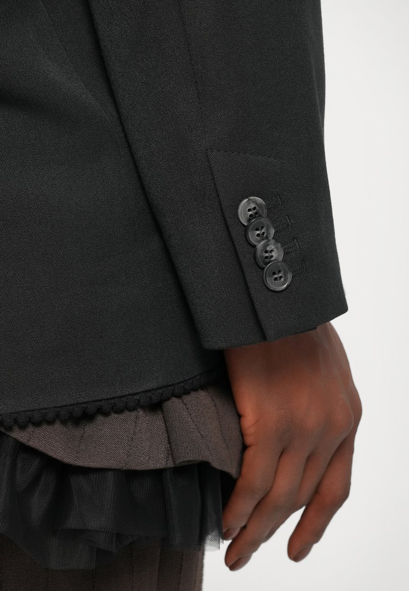 Schwarzer, maßgeschneiderter Blazer mit glatter Textur, ausgestattet mit vier schwarzen Knöpfen und einer Tasche. Darunter befinden sich stoffschichten aus dunkelbraunem Material und Tüll.