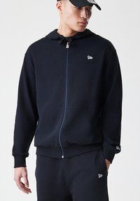 Hoodie zippé noir avec capuche à cordon, poches avant, poignets et ourlet côtelés, comportant un petit logo sur la poitrine. Coupe décontractée.
